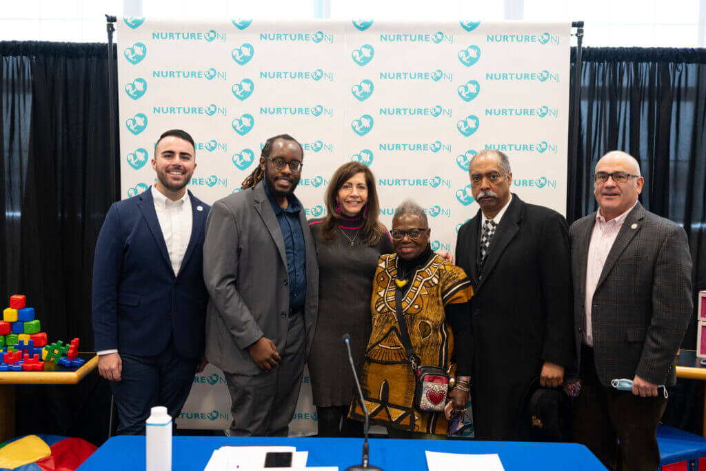 Members of the Nurture NJ Coalition — including NJ First Lady Tammy Murphy and Garden State Equality Executive Director Christian Fuscarino and Board Chair Jeannine Frisby LaRue — pose for a group photo after a press conference. They are in business formal attire, standing in front of a banner with the coalition's logo repeated as a pattern on it.